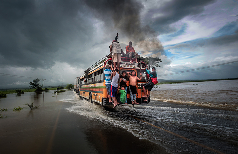 Ein Bus, auf dem sich hinten meherer Personen außen festhalten, fährt über eine überschwemmte Straße; am Himmel sind sehr dunkle Wolken zu sehen.